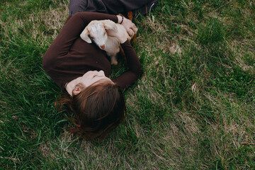Fototapeta premium Relaxed woman lying on grass, cuddling a goat kid in a sunny field, capturing a calm outdoor lifestyle moment of affection, companionship, and simple nature joy.