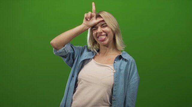 Young woman in denim shirt sticks tongue out and makes l hand sign on forehead in green screen studio; playful defiance.