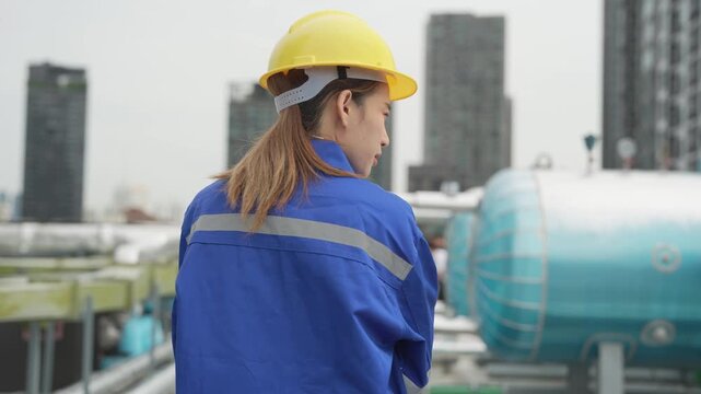 A female mechanical engineer in a yellow hardhat leans down to inspect industrial cooling tanks and pipe connections during a routine facility audit on a building rooftop.