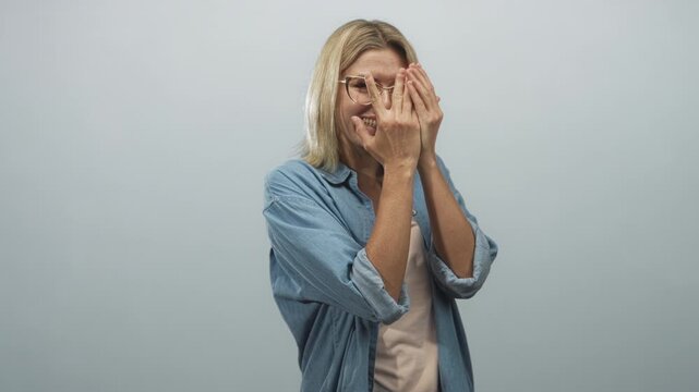 Woman covers face with hands, peeking between fingers, wearing glasses and denim shirt in studio; shyness vulnerability.