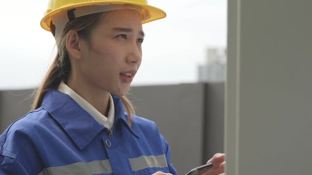 A female electrical engineer wearing a yellow hardhat and blue uniform operates a digital control panel display while holding a smartphone during a technical building system audit.