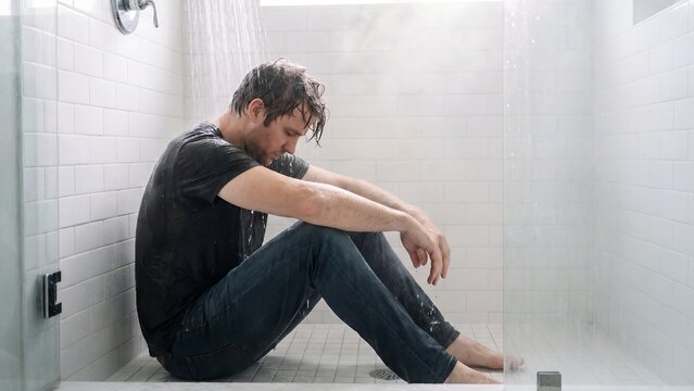 A man sits alone and dejected on the cold floor of a modern shower, head bowed in quiet despair, reflecting on personal struggles amidst the steamy bathroom