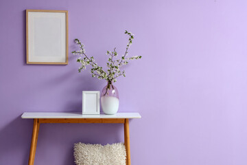 Fototapeta premium Vase with blooming tree branches and frame on table near lilac wall