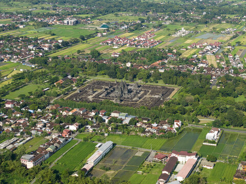 Aerial view of Sewu Temple complex surrounded by traditional houses, green rice fields, and tropical trees under bright daylight Kecamatan Prambanan, Daerah Istimewa Yogya, Indonesia.
