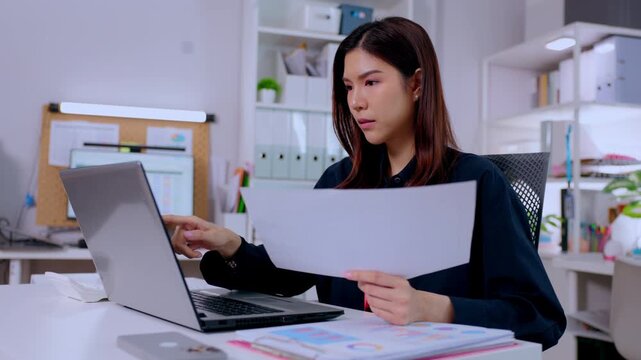 Focused Asian adult woman typing diligently on modern laptop computer at corporate office desk reviewing important business document paper while observing screen with serious concentrated expression