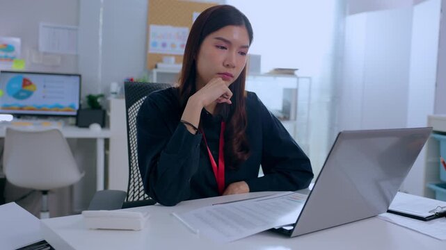 Serious asian business woman working late at modern office desk looking extremely focused thinking deeply reviewing complex financial document on laptop computer managing corporate project