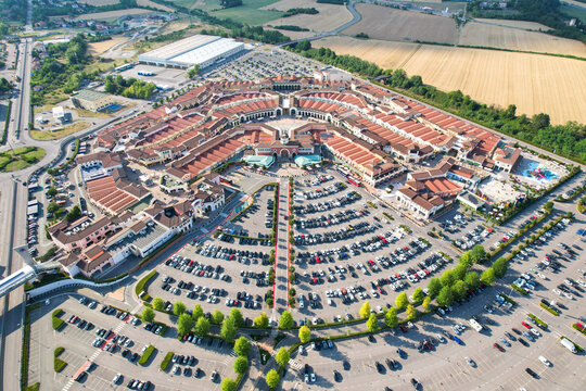 Aerial view of the Designer Outlet Serravalle featuring its circular architectural layout, red-tiled roofs, and large parking lots in Serravalle Scrivia, Piemonte, Italy.
