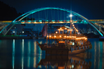 Fototapeta premium Motion blur night Seascape at the illuminated Gangguan Port with night cruise passing the arching bridge in Tongyeong‑si, Gyeongsangnam‑do, South Korea