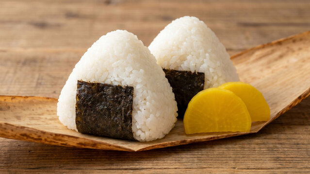 白いご飯のおにぎりと黄色いたくあんを写した、素朴で親しみやすい和食イメージ。海苔を巻いた三角おにぎりのつややかな米粒と木目の温もりが印象的で、朝食、軽食、弁当、家庭料理、日本食、和の食卓、飲食店メニュー、食品広告に適したナチュラルで清潔感のあるフード写真