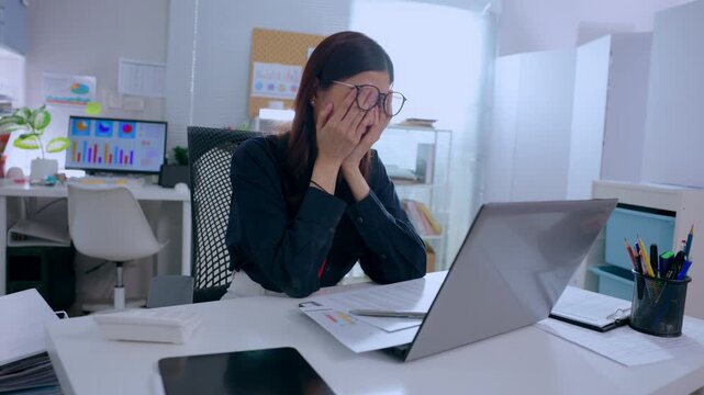 Stressed businesswoman working late at modern corporate office desk typing document on laptop feeling exhausted adjusting glasses covering face with hands experiencing serious burnout