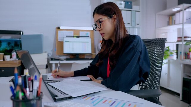 Serious businesswoman focused on writing financial document at modern dark office desk working late night overtime feeling tired yet determined analyzing project schedule near laptop computer screen