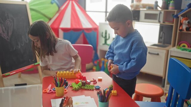 Girl and boy stacking colorful plastic blocks and sorting crayons on a red table inside a building playroom; focused play.