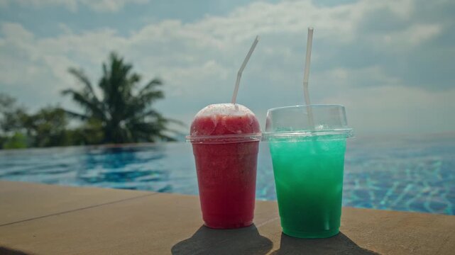 Two colorful frozen fruit cocktails standing near infinity swimming pool with palm tree background during sunny summer vacation.