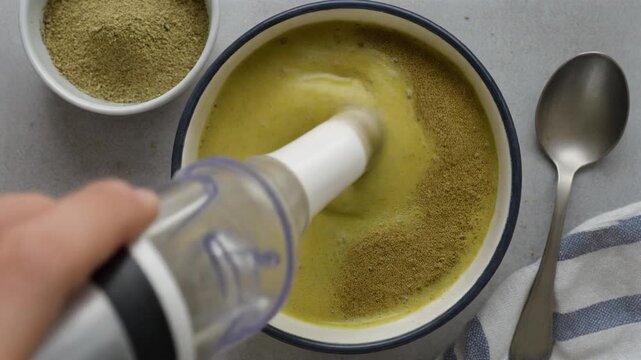 A person blending a green drink in a bowl with an immersion blender, with green powder nearby.