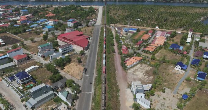 Forward aerial view of a long freight train crossing a town intersection beside an extensive palm plantation in Kampot
