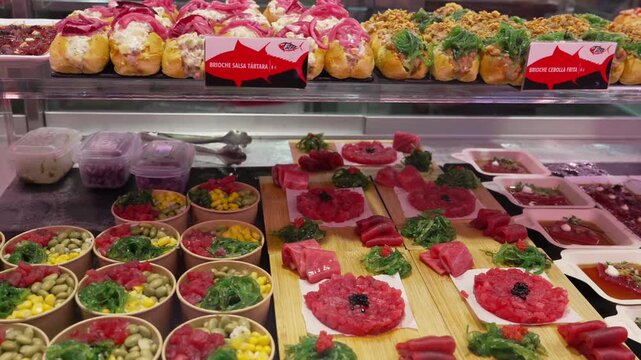 Assorted tapas at Mercado de San Miguel Madrid Spain include tuna tartare Thunnus sp with herbs and vegetables alongside prepared salads and filled appetizers in deli display, slow pan right