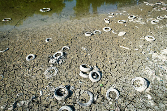 Aerial view of discarded tires scattered across a cracked and parched riverbed under bright sunlight in Carignano, Piemonte, Italy.