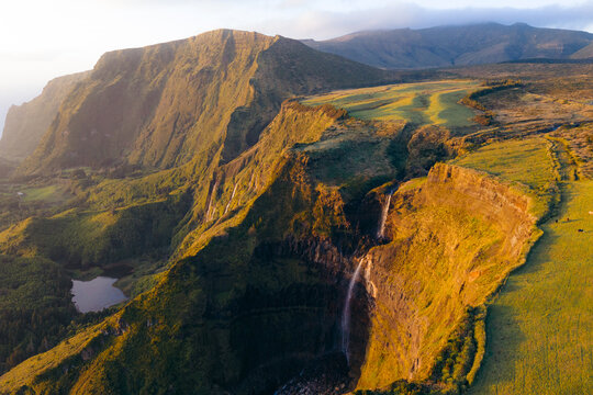 Aerial view of Poco da Ribeira do Ferreiro waterfalls cascading down steep green cliffs during golden hour in Fajazinha, Azores, Portugal.