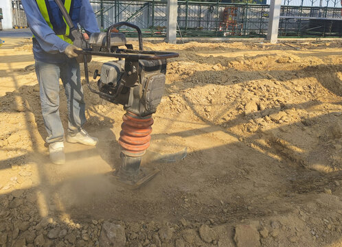 A construction worker is using a jumping jack compactor to compact soil in a factory area for construction site preparation.