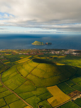 Aerial view of Pico Dona Joana geodesico volcanic crater surrounded by green agricultural fields and stone walls overlooking the Atlantic Ocean in Feteira, Acores, Portugal.
