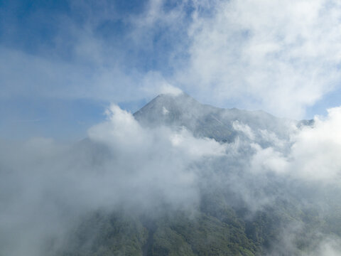 Aerial view of the majestic peak of Mount Merapi partially covered by thick white clouds under a clear blue sky in Kecamatan Cangkringan, Daerah Istimewa Yogya, Indonesia.