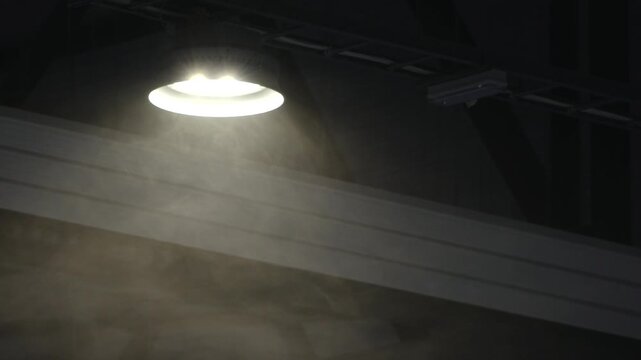 Bright ceiling lamp shines through dust and mist in a dark interior, creating a moody atmosphere with dramatic light beams and deep shadows.