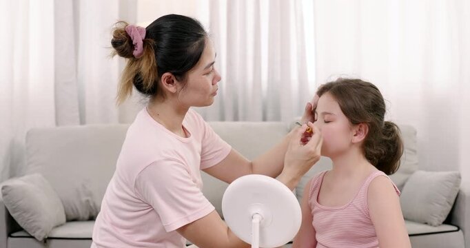 Asian adult female focuses while applying mascara to eyelashes of multiracial girl aged 8 seated quietly on sofa during bonding beauty lesson in home environment with care love and soft parenting