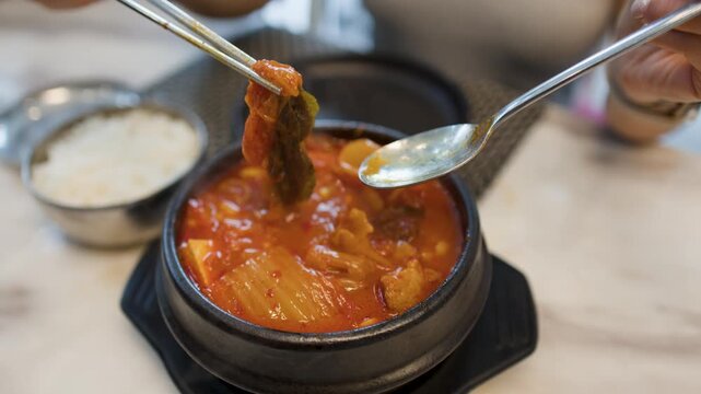 Person Eating Spicy Korean Kimchi Jjigae Stew with Metal Chopsticks