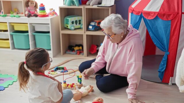 Woman in pink hoodie and glasses hands a doll to a little girl, both seated on the floor among toys, storage bins and a play tent in a playroom building; tenderness.