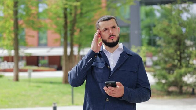 Individual with headache holding phone outdoors. Stress and discomfort apparent on face. Modern life challenges creating physical strain. Feeling worried and pensive amidst green surroundings.