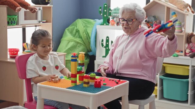 Senior woman and little girl play with plastic blocks at a small table, woman lifts a multicolored toy while the child stacks pieces in a building playroom; intergenerational bonding play.