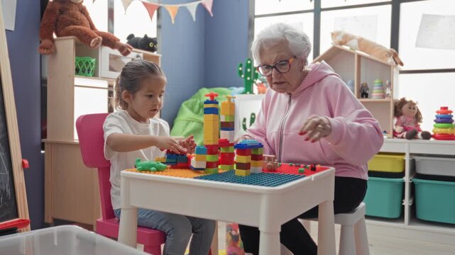 Senior woman and little girl build colorful block tower on a small table in a building playroom filled with toys; warmth and bonding.