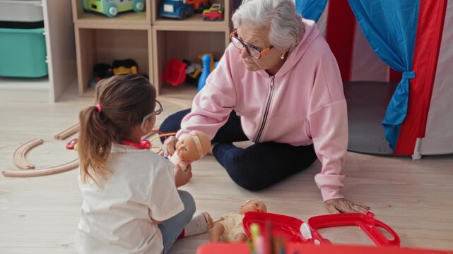 Senior woman holds baby doll while watching little girl play doctor with toy stethoscope in building; intergenerational tenderness.