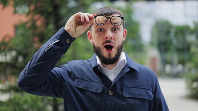 Man expresses deep surprise and disbelief, lifting glasses and widening eyes. Astonishment and wonder are clearly visible on face as reacts with open mouth and raised eyebrows.