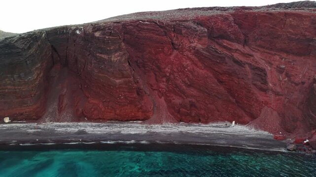 Aerial view of Red beach featuring steep red volcanic cliffs and dark pebbled shoreline under soft light in Fira, Greece.