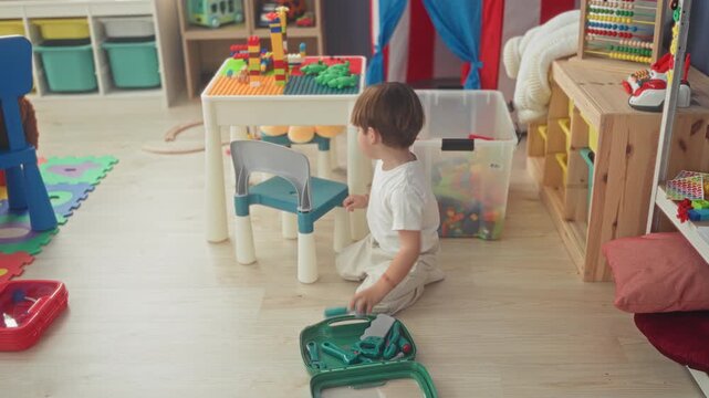 Boy kneeling playing with green toy toolkit on floor in building; curiosity imagination learning discovery.