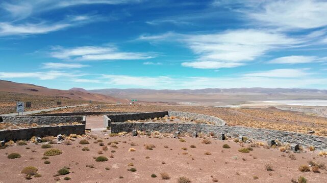 Panoramic aerial shot of abandoned stone border checkpoint ruins on high altitude plateau with salt lake and vast Andean mountain range under blue sky. Paso Jama, Chile