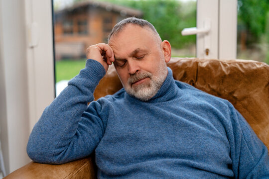 Man sits on couch in a cozy room with eyes closed and hand on forehead while resting in the afternoon