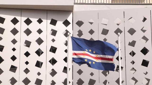 The national flag of Cabo Verde waving on a flagpole in front of a modern white building featuring a contemporary geometric diamond-patterned facade