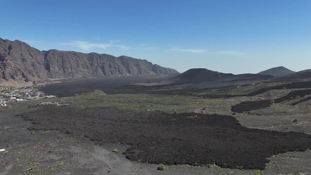 Aerial wide shot of the vast, black solidified lava fields in the Ch&atilde; das Caldeiras basin, set against the towering Bordeira cliff wall on Fogo Island, Cabo Verde