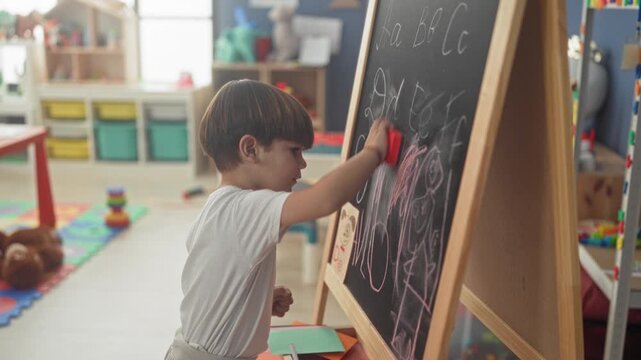 Little boy draws with chalk on a blackboard easel inside a classroom building, reaching for chalk and eraser on the tray; quiet concentration.
