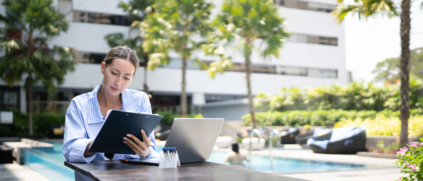 Professional businesswoman reviewing clipboard with laptop at luxury hotel poolside outdoor workspace