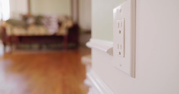 A hand inserts and removes then re-inserts black electrical plug into wall outlet with blurred living room in background dynamic shot.