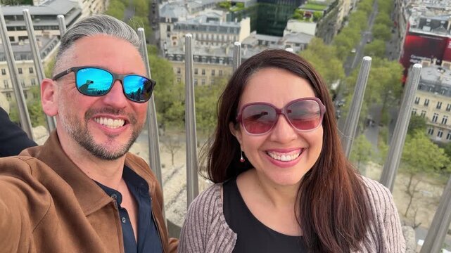 Joyful couple with sunglasses taking a selfie video from a viewpoint overlooking the Paris skyline, with the man pointing at the iconic Eiffel Tower in the background during their european vacation