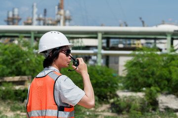 Obraz premium Asian male technician speaking by walkie talkie near refinery pipeline route, relaying petrochemical field update for maintenance planning, safety audit, hazard control and operational monitoring