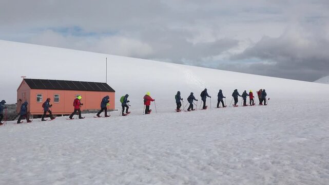 A group of people in bright jackets walk together over the vast white snow toward a tall mountain. The sky is gray and cloudy as they make their way through Antarctica.