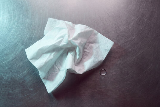 Remnants of release. A crumpled white tissue and a single tear drop on a brushed metal surface. Minimalist symbolic representation of healing and closure.

