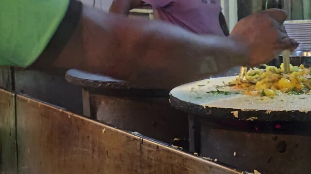 A skilled street food cook smashes seasoned potato filling onto a thin, crispy dosa cooking on a hot tawa pan. The flavorful masala is spread wide before the iconic South Indian crepe is served hot. 