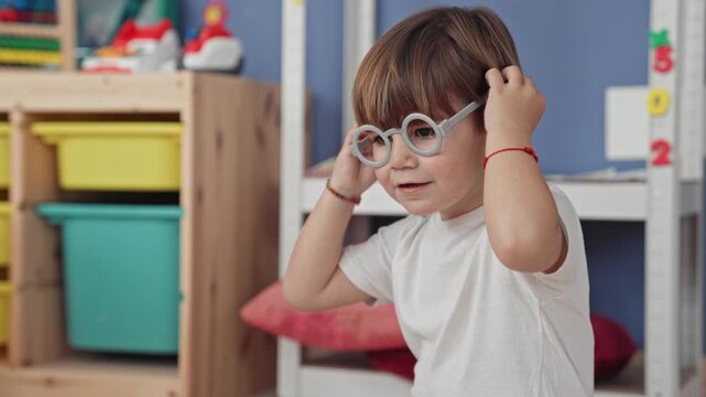 Boy in white tshirt puts on round toy glasses and smiles, holding frames with his hand while seated among shelves and bins in a studio; playful joy.
