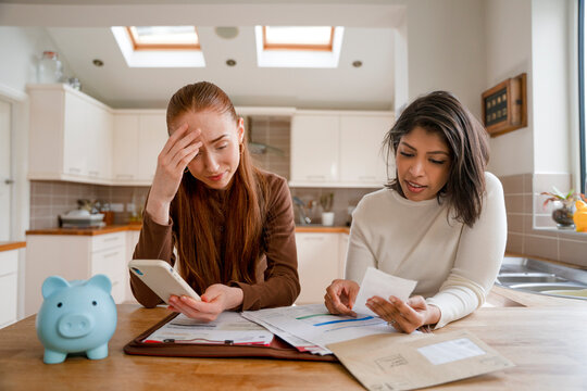 Two women discuss bills and finances at a kitchen table while using a phone and reviewing documents during the day